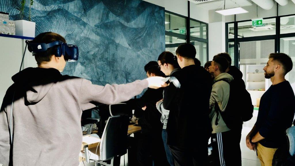 Grupa młodzieży stojącej tyłem, w nowoczesnym wnętrzu, na pierwszym planie chłopak z założonymi okularami VR i kontrolerem trzymanym w dłoni