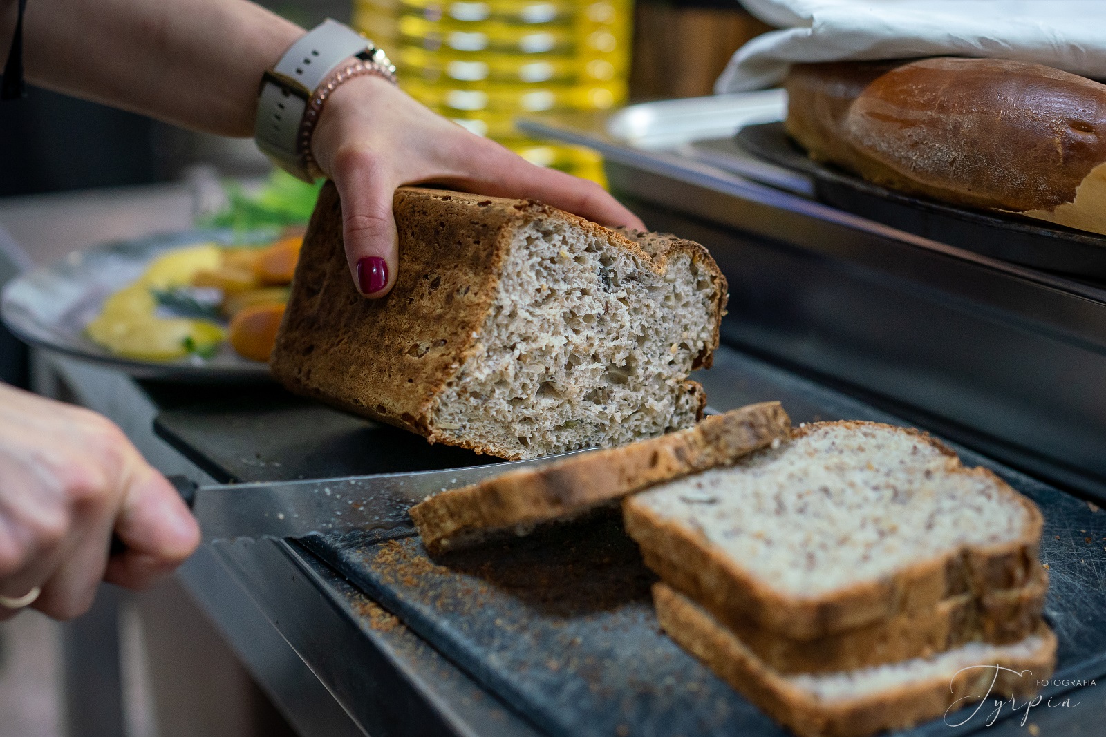 Chleb razowy na czarnej desce, trzymany przez kobiece dłonie w trakcie krojenia.