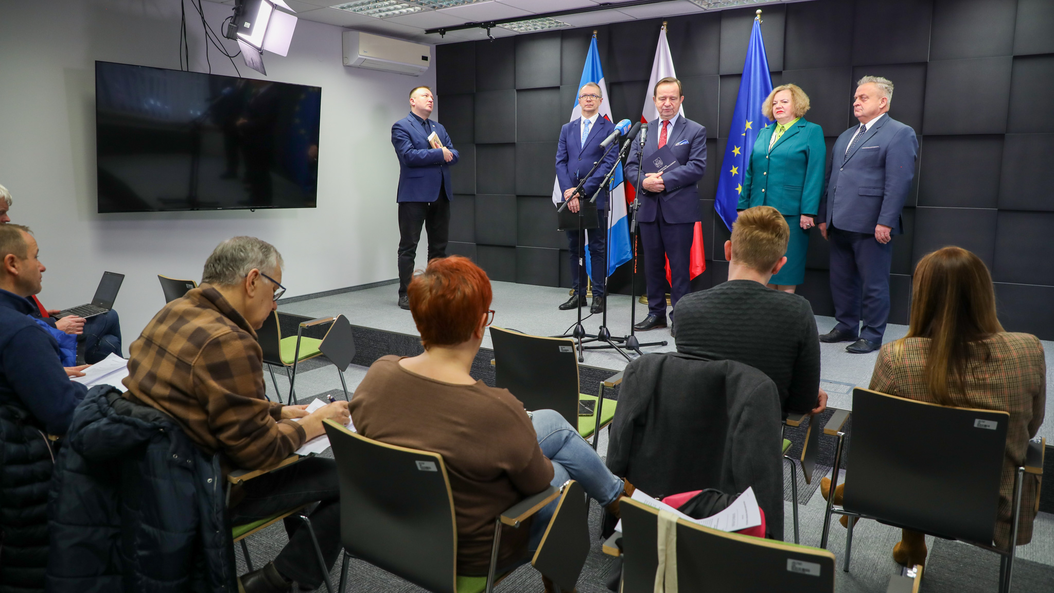 Na scenie na tle czarnej ściany i flag województw apodkarpackiego, RP i UE stoi pięć osób elegancko ubranych: jedna kobieta i czterech mężczyzn. Przed nimi siedzą dziennikarze, tyłem do fotografa.